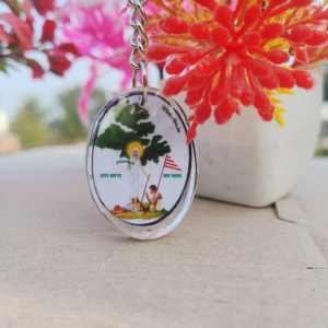 An elegant Sarna Maa oval resin keychain measuring 4 cm x 3 cm, featuring a detailed illustration of Sarna Maa standing under a sacred tree with the text "Jai Sarna" and "Jai Johar." The keychain is lightweight, durable, and perfect for celebrating tribal culture and spirituality. Captured with vibrant decorative plants in the background, this handmade keychain symbolizes Jharkhand's rich tribal heritage.