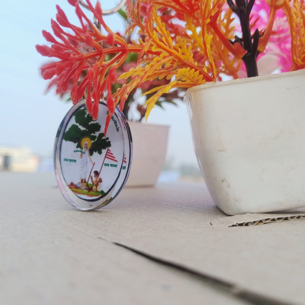 An elegant Sarna Maa oval resin keychain measuring 4 cm x 3 cm, featuring a detailed illustration of Sarna Maa standing under a sacred tree with the text "Jai Sarna" and "Jai Johar." The keychain is lightweight, durable, and perfect for celebrating tribal culture and spirituality. Captured with vibrant decorative plants in the background, this handmade keychain symbolizes Jharkhand's rich tribal heritage.