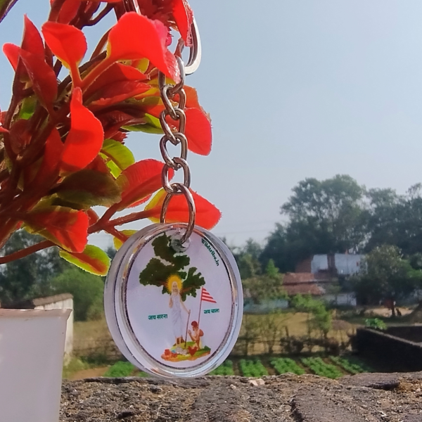 A beautifully crafted Sarna Maa resin keychain hanging next to a vibrant red plant in a white pot. The round keychain, measuring 2 cm in diameter, features an intricate illustration of Sarna Maa, symbolizing tribal culture and spirituality. Perfect for gifting or daily use, this handmade accessory celebrates the rich heritage of Jharkhand.