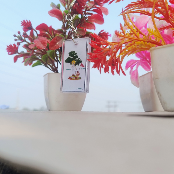 An elegant Sarna Maa oval resin keychain measuring 4 cm x 3 cm, featuring a detailed illustration of Sarna Maa standing under a sacred tree with the text "Jai Sarna" and "Jai Johar." The keychain is lightweight, durable, and perfect for celebrating tribal culture and spirituality. Captured with vibrant decorative plants in the background, this handmade keychain symbolizes Jharkhand's rich tribal heritage.