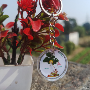 A beautifully crafted Sarna Maa resin keychain hanging next to a vibrant red plant in a white pot. The round keychain, measuring 2 cm in diameter, features an intricate illustration of Sarna Maa, symbolizing tribal culture and spirituality. Perfect for gifting or daily use, this handmade accessory celebrates the rich heritage of Jharkhand.