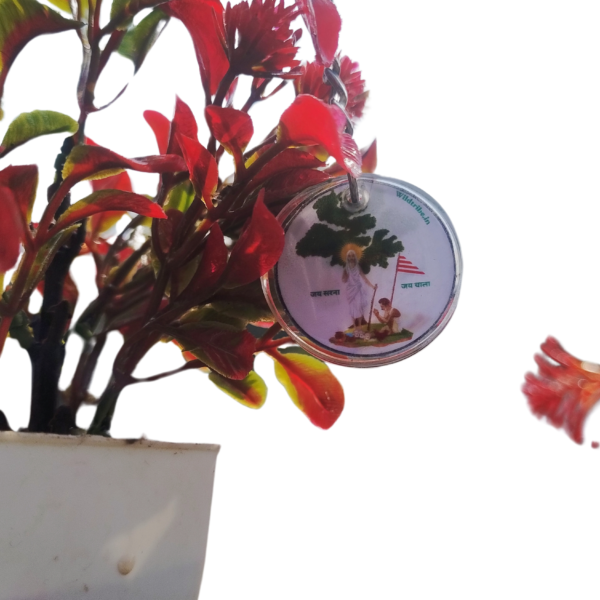 A beautifully crafted Sarna Maa resin keychain hanging next to a vibrant red plant in a white pot. The round keychain, measuring 2 cm in diameter, features an intricate illustration of Sarna Maa, symbolizing tribal culture and spirituality. Perfect for gifting or daily use, this handmade accessory celebrates the rich heritage of Jharkhand.